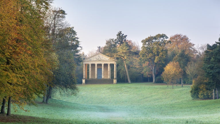 A view across misty parkland surrounded by trees towards the Grecian Valley at Stowe, Buckinghamshire.